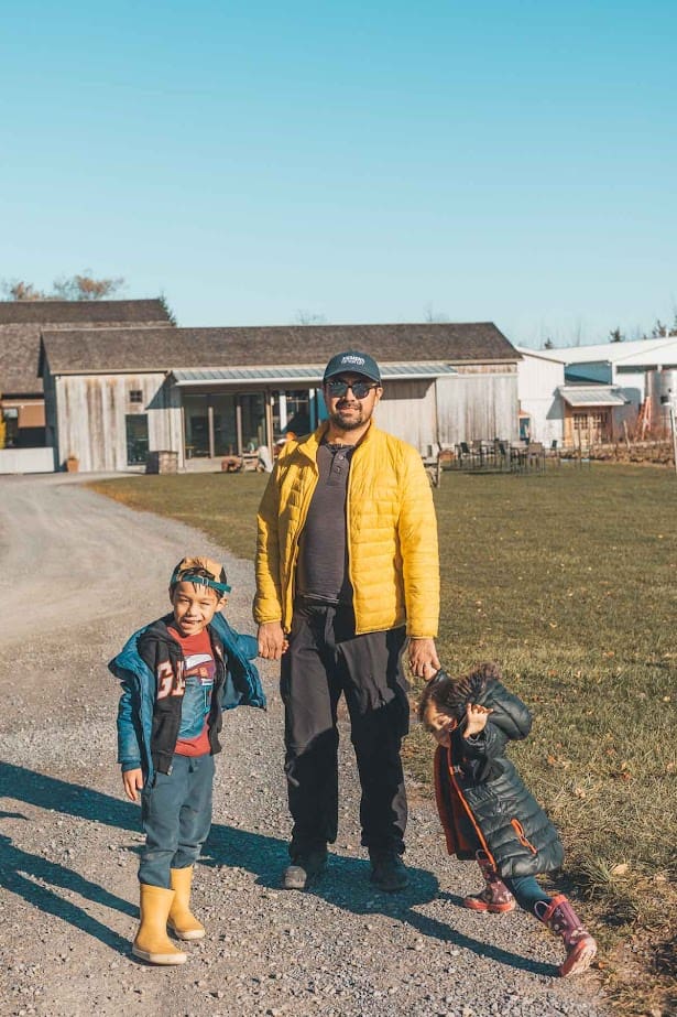 A man in a yellow jacket stands outside with two young children on a sunny day near farm buildings, reminiscent of the scenic landscapes found at Finger Lakes Wineries with Lodging.