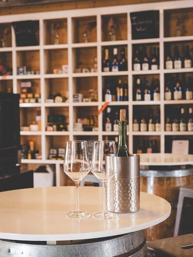 Wine glasses and a bottle in a bucket on a round table, set against shelves of wine bottles—evoking the charm of the best wineries in Finger Lakes.
