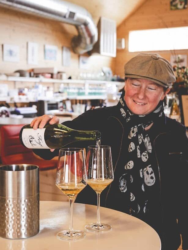 A person in a cap pours white wine into two glasses at a table in a cozy, well-lit café or wine bar—reminiscent of the inviting ambiance found at the best wineries in Finger Lakes.