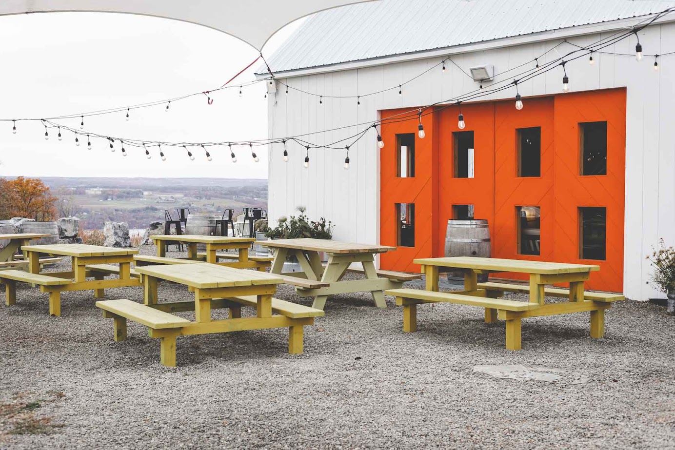 Outdoor picnic tables on gravel near a white building with bright orange doors and hanging string lights, evoking the relaxed charm found at the best wineries in Finger Lakes.