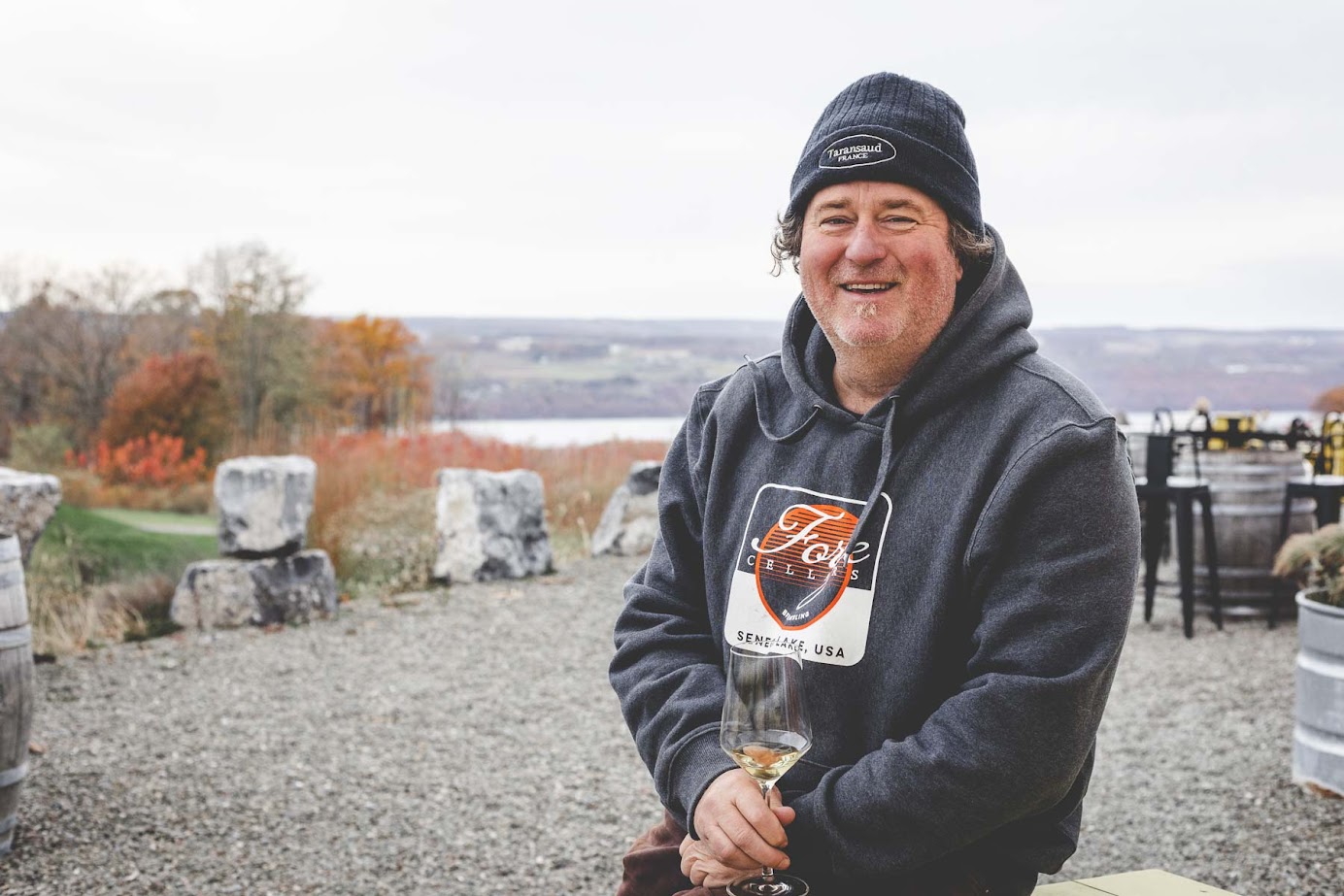 Smiling man in a beanie and hoodie holding a wine glass outdoors, enjoying autumn scenery at one of the best wineries in Finger Lakes.