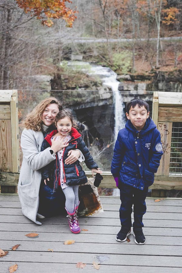 A woman and two children pose on a wooden deck at Watkins Glen State Park, with a waterfall in the background on an autumn day.