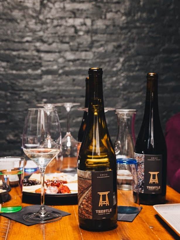 Two wine bottles and glasses on a wooden table with food, set against a dark brick wall background—perfect for savoring selections from the best wineries in Finger Lakes.