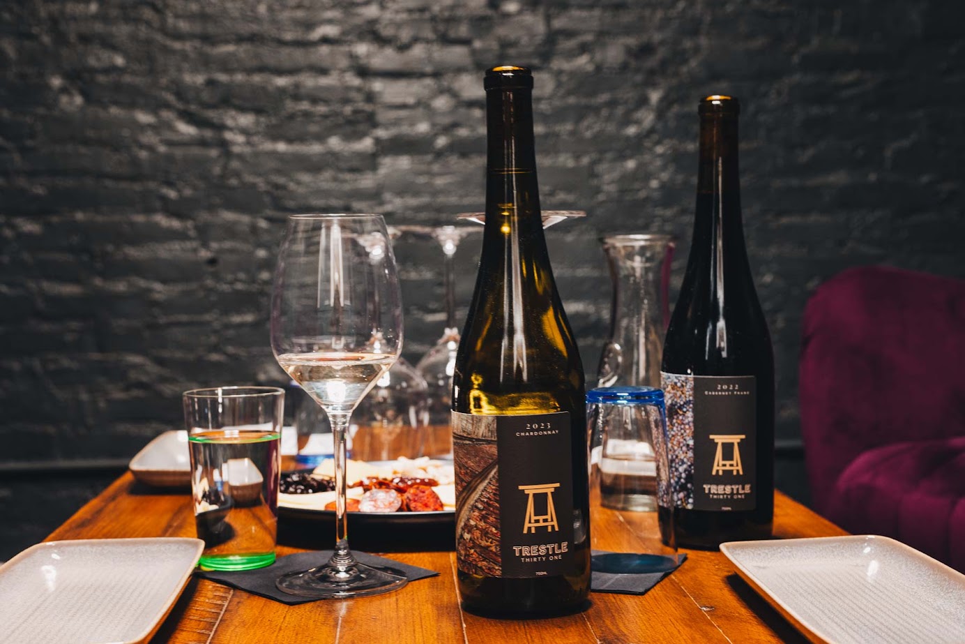 Two wine bottles, glasses, and plates are set on a wooden table against a dark brick wall background, evoking the ambiance of the best wineries in Finger Lakes.