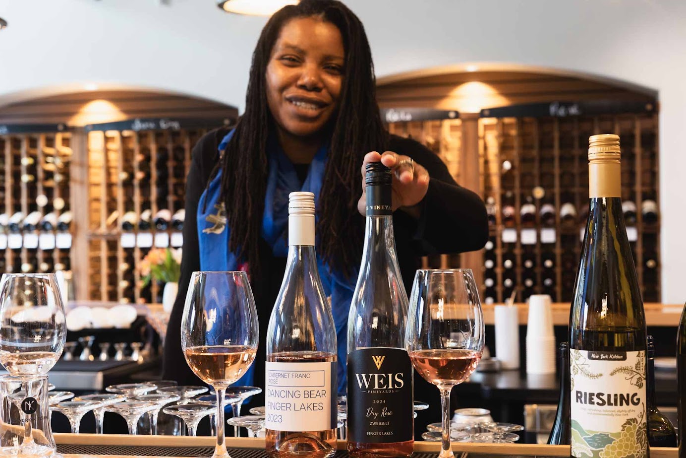 Smiling woman stands behind a bar with wine bottles and glasses in a wine-tasting room at one of the best wineries in Finger Lakes.