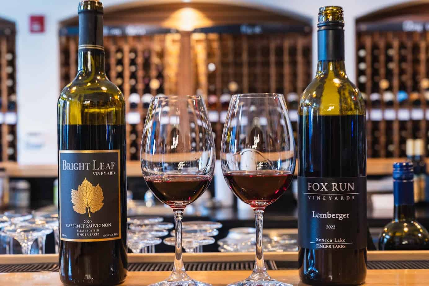 Two glasses of red wine and two wine bottles displayed on a bar with a wine rack in the background—perfect for a Finger Lakes weekend getaway.