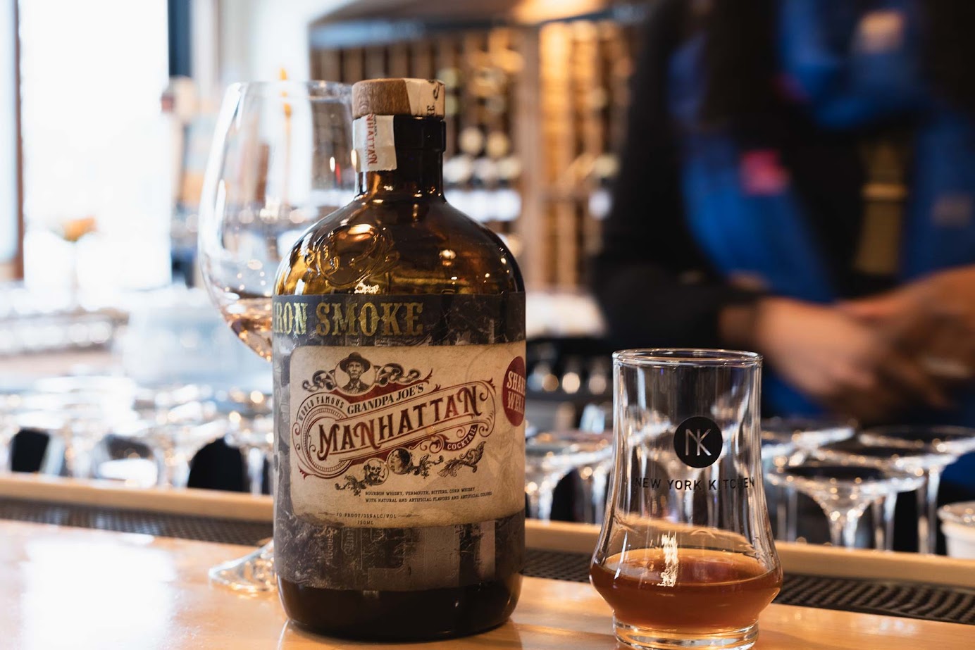 A bottle of Manhattan cocktail and a glass on a bar counter, evoking the elegance found at the best wineries in Finger Lakes, with blurred background and glassware.