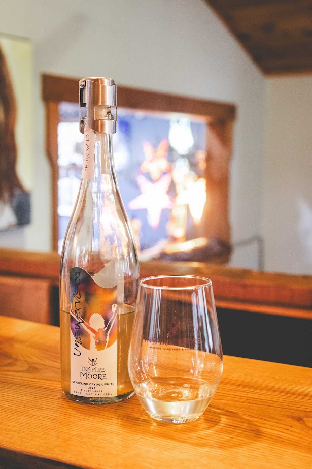A bottle of Inspire Moore wine and a glass with white wine on a wooden counter indoors, showcasing the craftsmanship found at one of the best wineries in Finger Lakes.