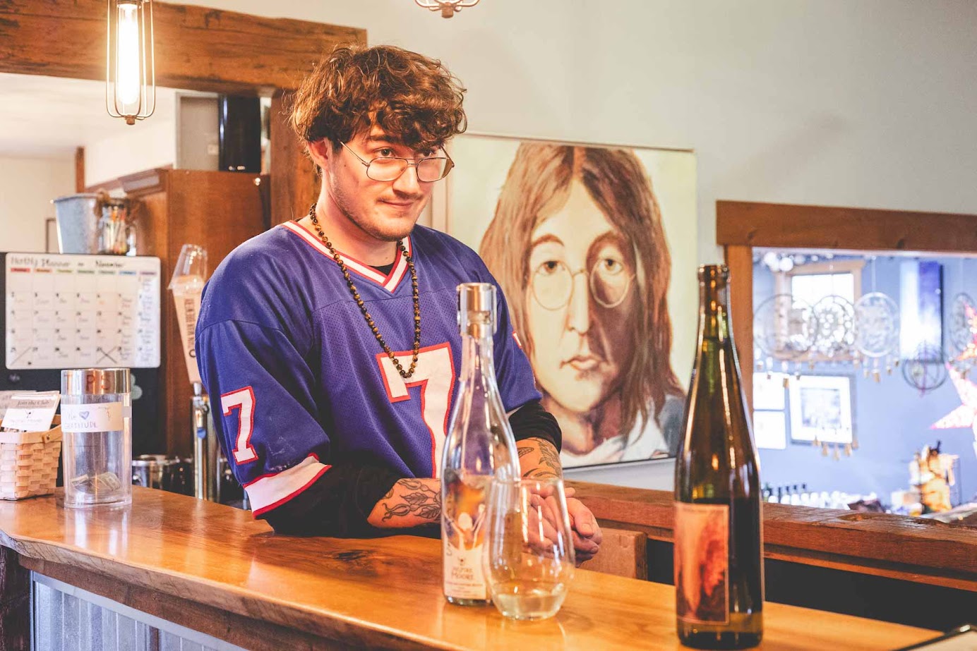 A man in a blue jersey stands behind a bar with bottles and glasses, evoking the inviting atmosphere of the best wineries in Finger Lakes, with a portrait on the wall behind him.