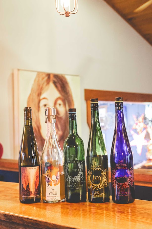 Five assorted wine and spirit bottles from the best wineries in Finger Lakes rest on a wooden counter, with artwork and a fish tank in the background.