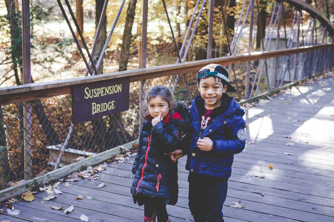 Two kids in jackets stand smiling on a wooden bridge with a "Suspension Bridge" sign in the forest at Watkins Glen State Park.