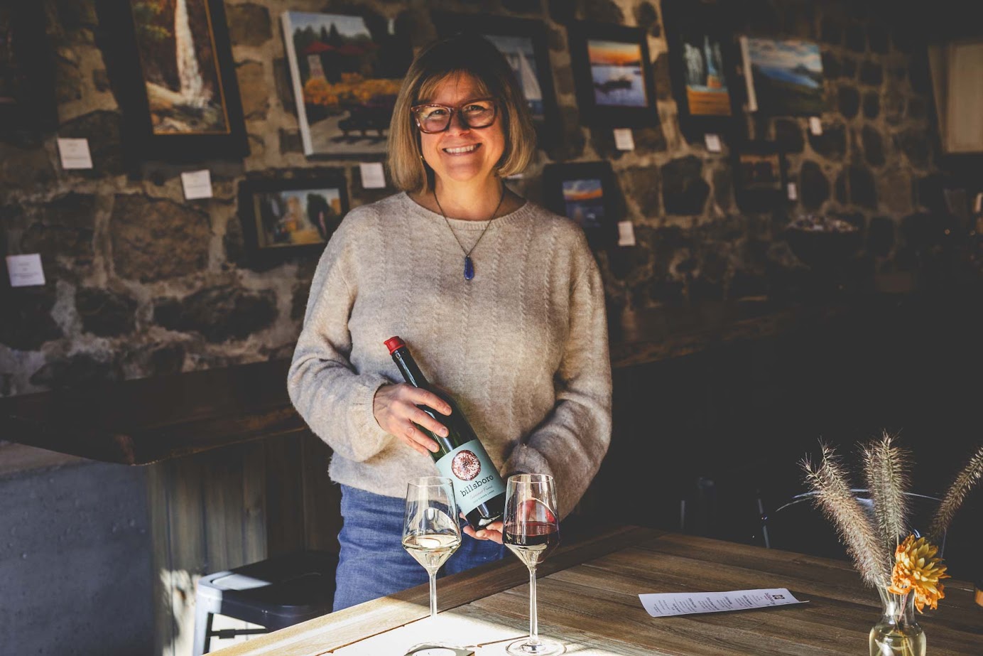 Smiling woman holding a wine bottle beside two filled glasses in a cozy, art-filled room with stone walls—an inviting scene reminiscent of the best wineries in Finger Lakes.
