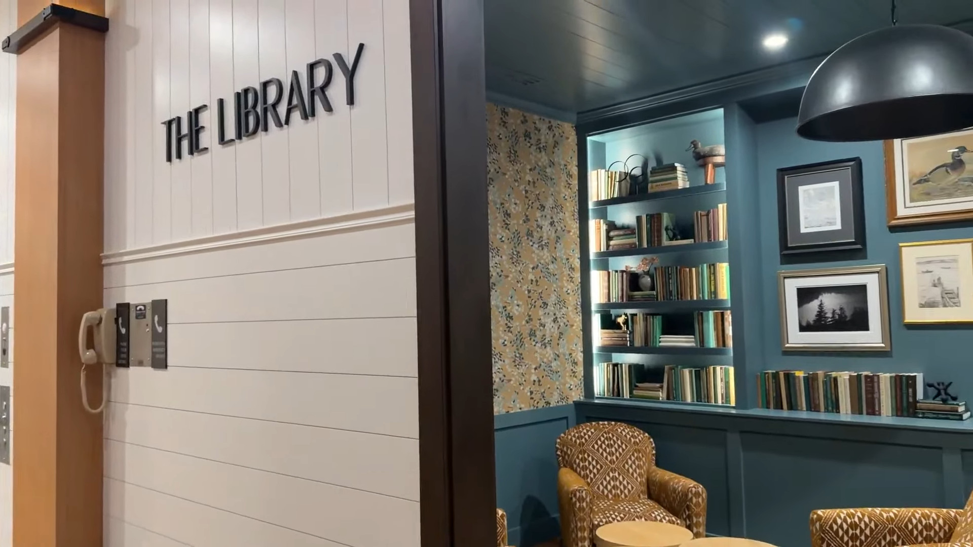A cozy library at Skaneateles Fields Resort with blue bookshelves, framed pictures, and a patterned armchair by a small table—a peaceful retreat after a relaxing visit to the spa in the scenic Finger Lakes.