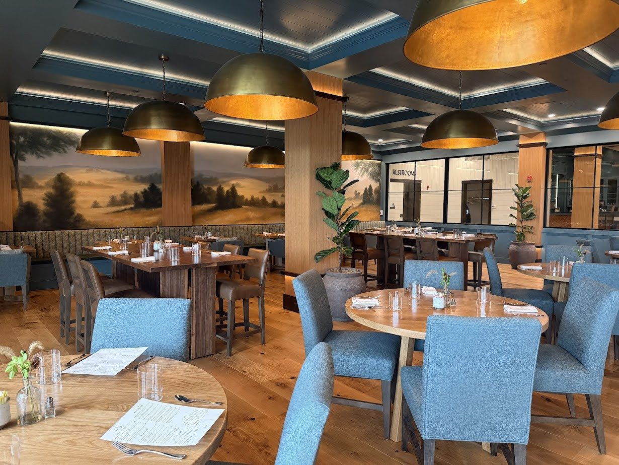 Modern restaurant with wooden tables, blue chairs, large gold pendant lights, and a mural of a Finger Lakes countryside landscape—perfect for a weekend getaway or fall road trip escape.