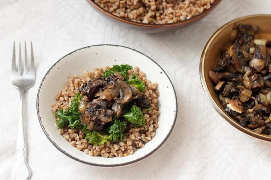 A bowl of buckwheat topped with saut&eacute;ed mushrooms and greens, a classic example of vegetarian dishes in Russia, with a fork and extra mushrooms beside it.