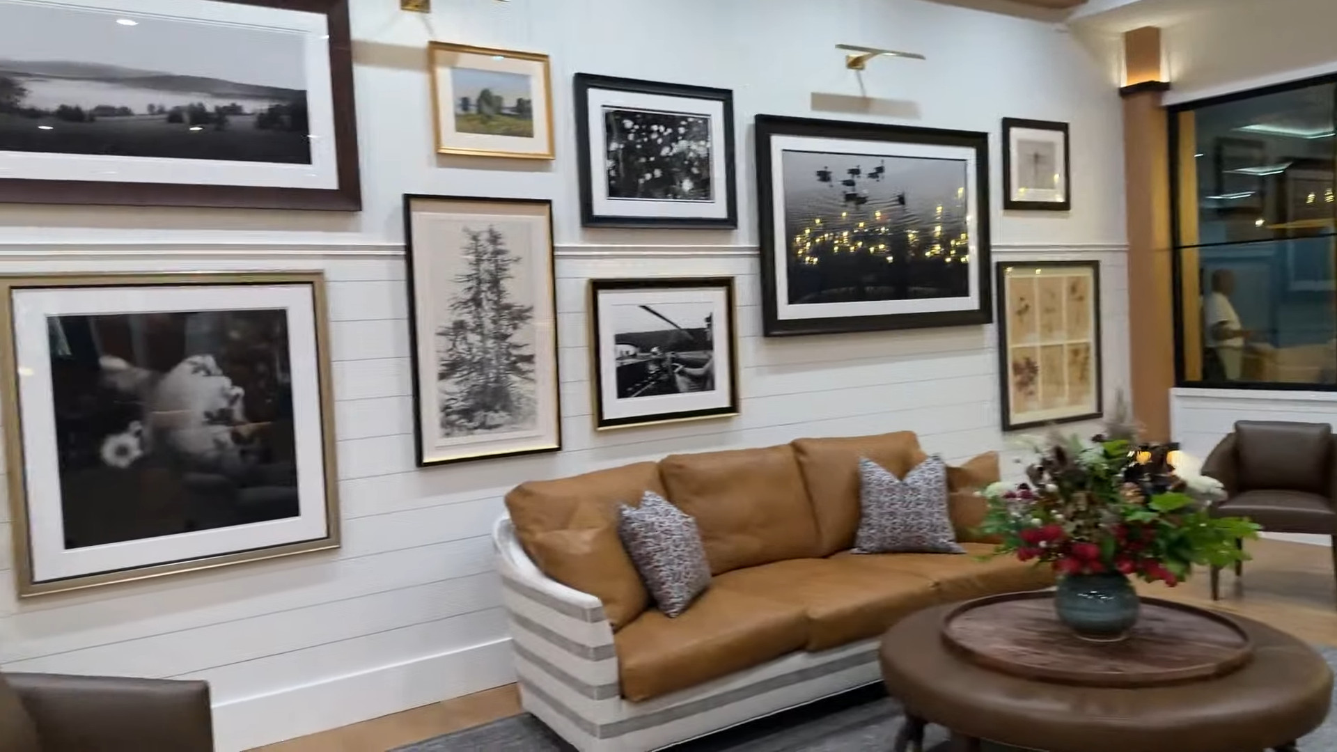 Modern living room with tan leather sofa, art-filled gallery wall, and round table with a floral centerpiece—creating a tranquil vibe that echoes the relaxing ambiance of Skaneateles Fields Resort & Spa in the Finger Lakes.