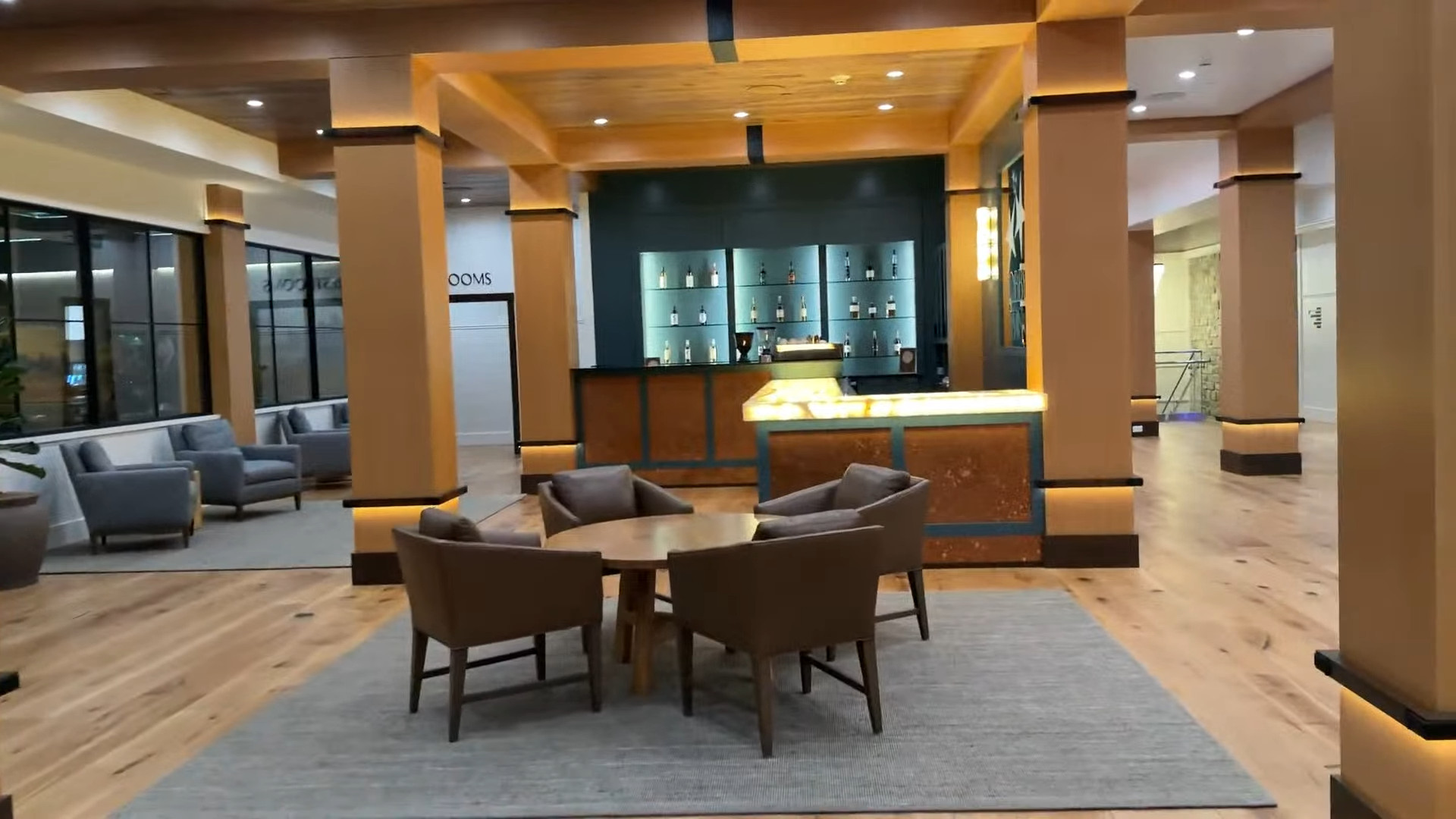 Modern lounge with a round table, four chairs, wooden floors, and a bar in the background—perfect for relaxing after exploring the Finger Lakes or visiting Spa at Skaneateles Fields Resort.