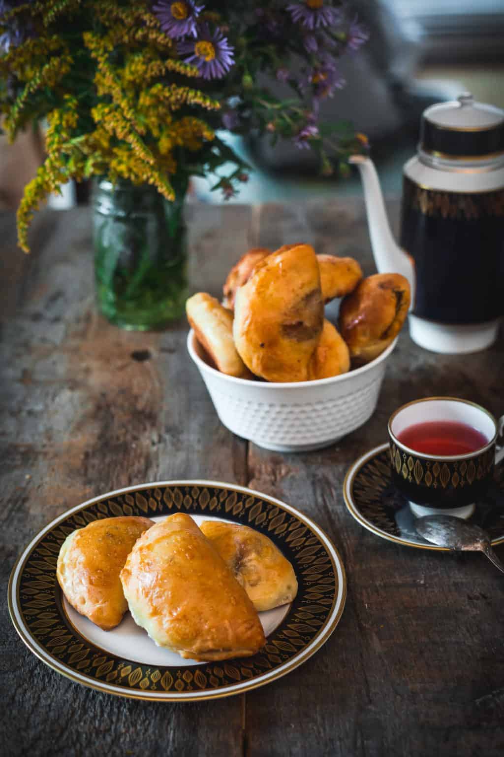 Two baked pastries on a plate, a cup of tea, and a bowl of vegetarian dishes in Russia are set on a rustic wooden table with flowers.