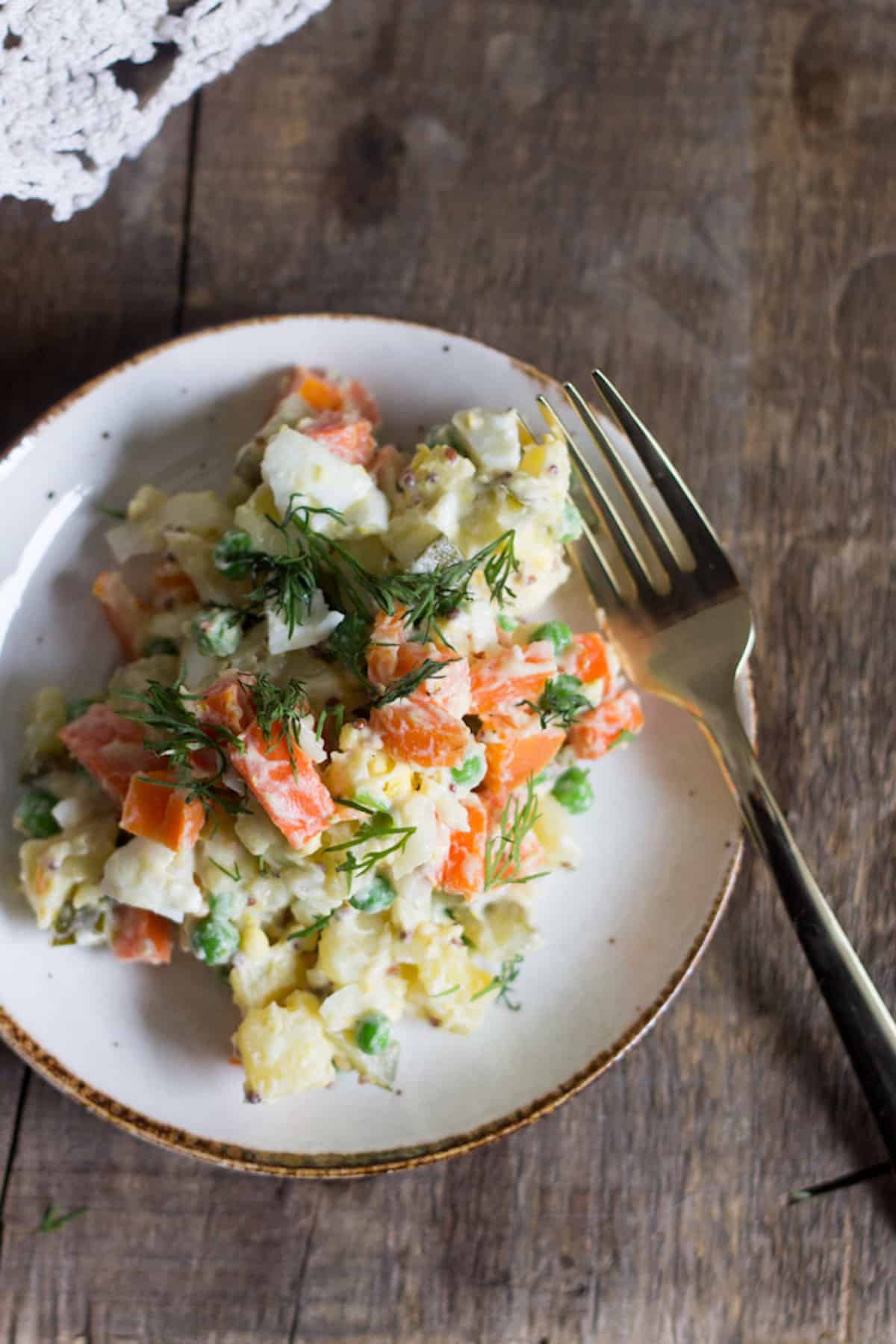 A plate of vegetable salad with carrots, peas, potatoes, and dill&mdash;one of the popular vegetarian dishes in Russia&mdash;served on a rustic wooden table with a fork.