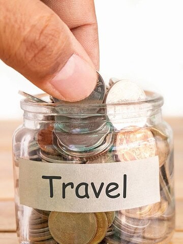 A hand puts coins into a glass jar labeled "Travel" with a passport and card in the background.