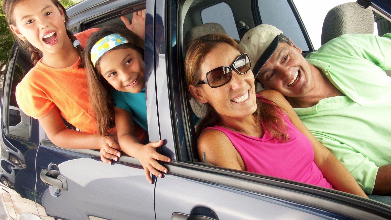 A smiling family of four leans out of a car window, ready for a trip.