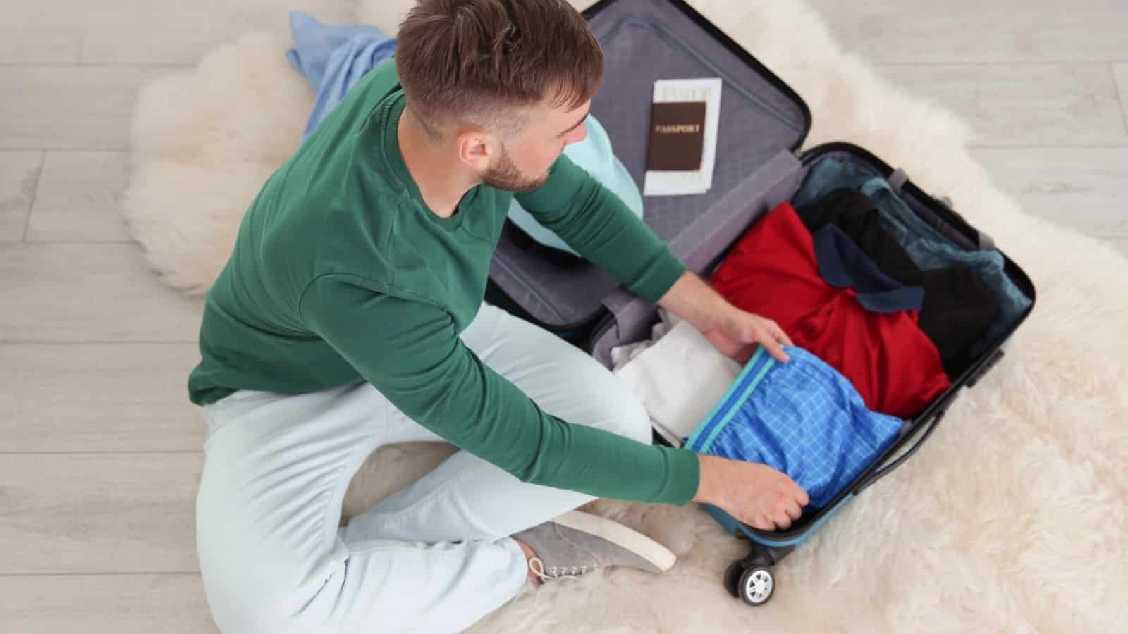 Man sitting on floor packing a suitcase with clothes and a passport visible inside the luggage.