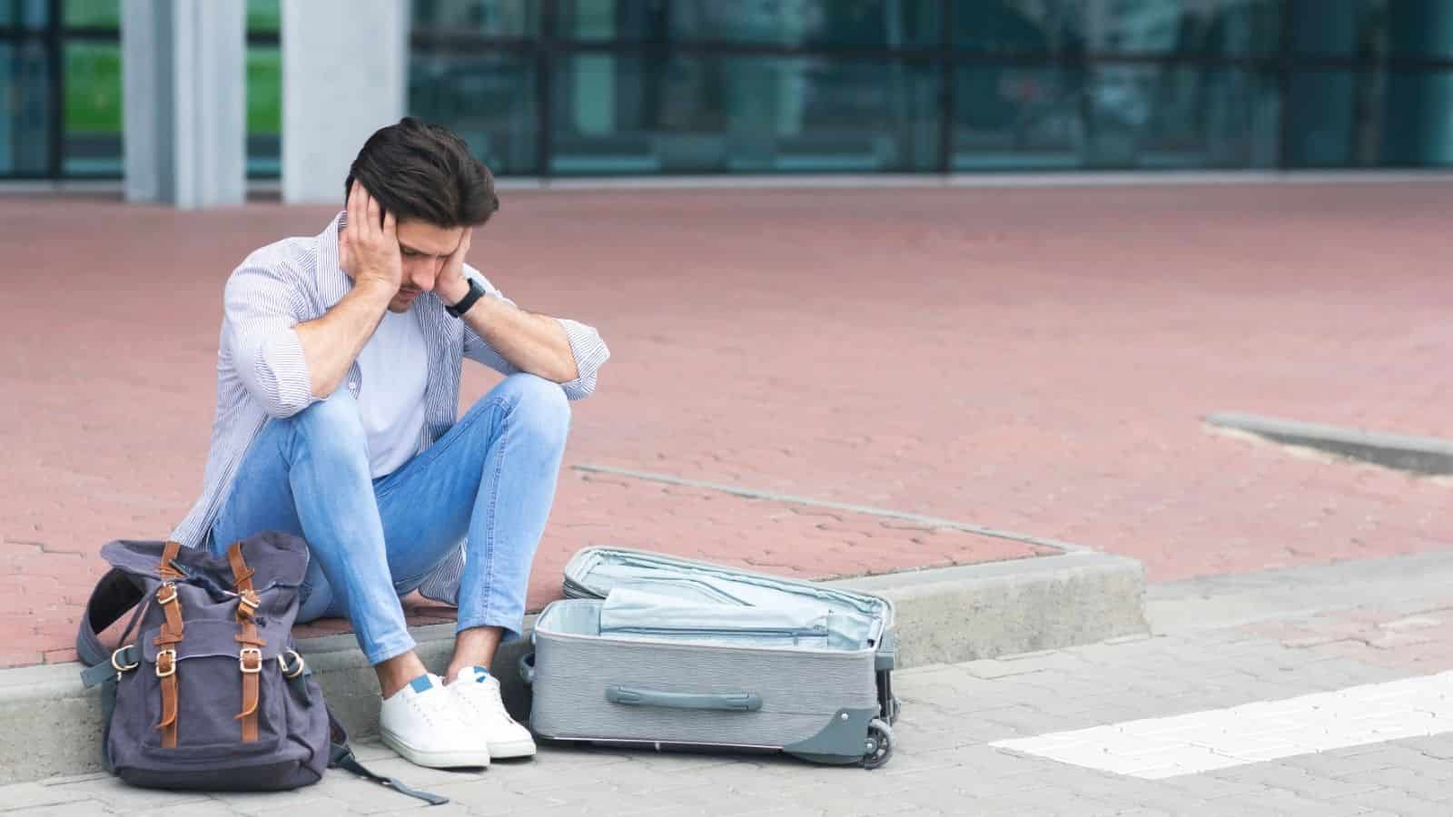 A man sits on a curb, looking stressed, with an open suitcase and backpack beside him.
