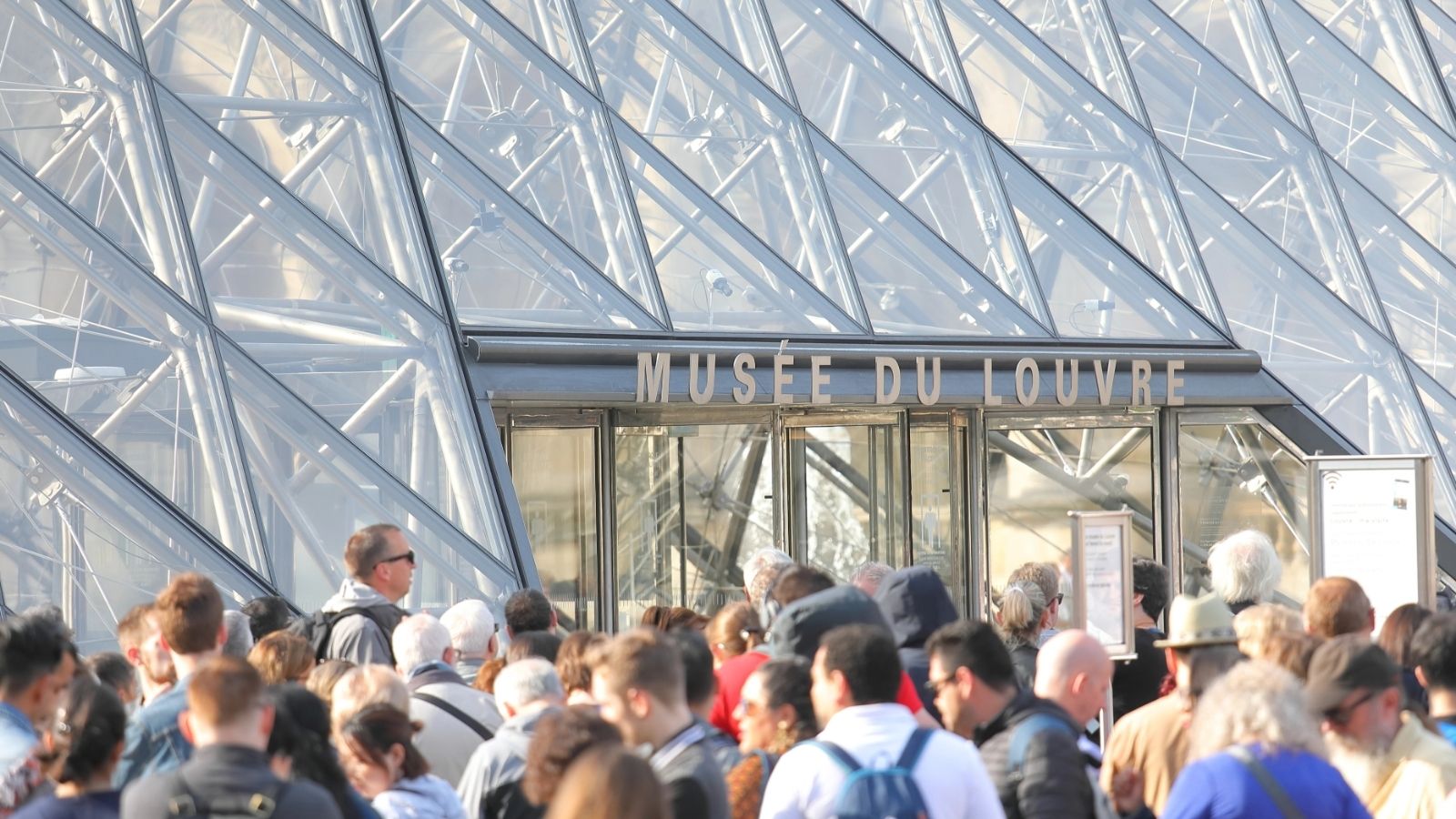 A crowd gathers at the glass entrance of the Mus&eacute;e du Louvre in Paris on a sunny day.