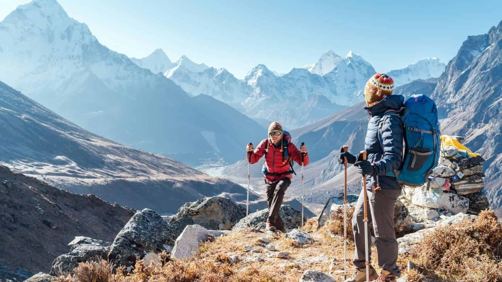 Two hikers trekking through rugged Himalayan mountain terrain with snow-capped peaks in the backgrou.