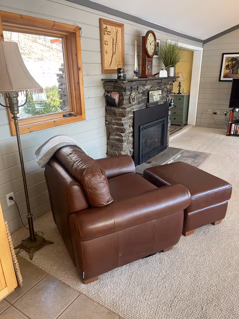 A brown leather armchair with ottoman sits by a stone fireplace in a cozy, sunlit living room, evoking the warmth and comfort of classic cottages and cabins—perfect for relaxing during Finger Lakes vacation rentals.