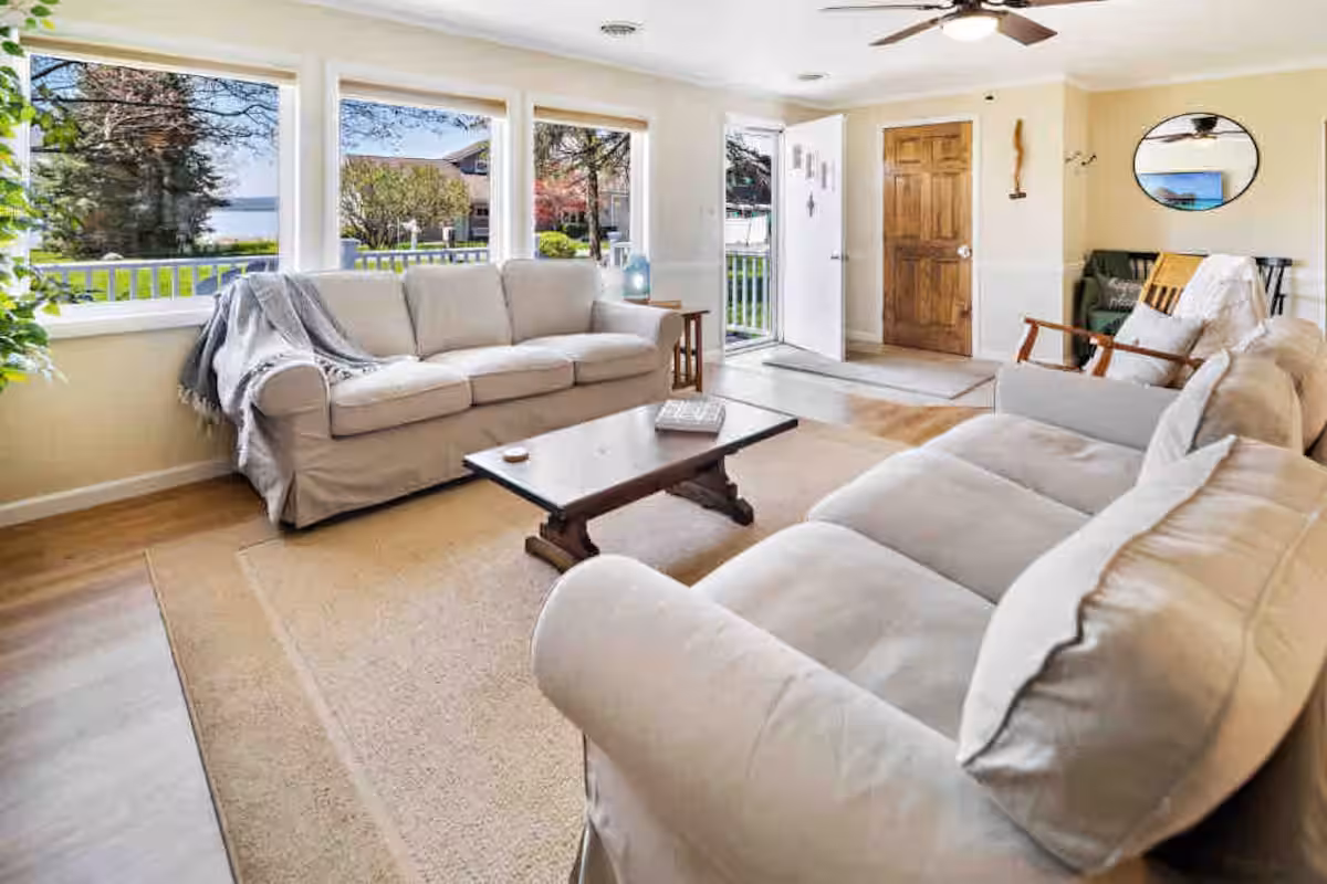 Bright living room with two beige sofas, a coffee table, large windows, and a wooden front door—perfect for relaxing in cozy Finger Lakes vacation rentals or charming cottages.