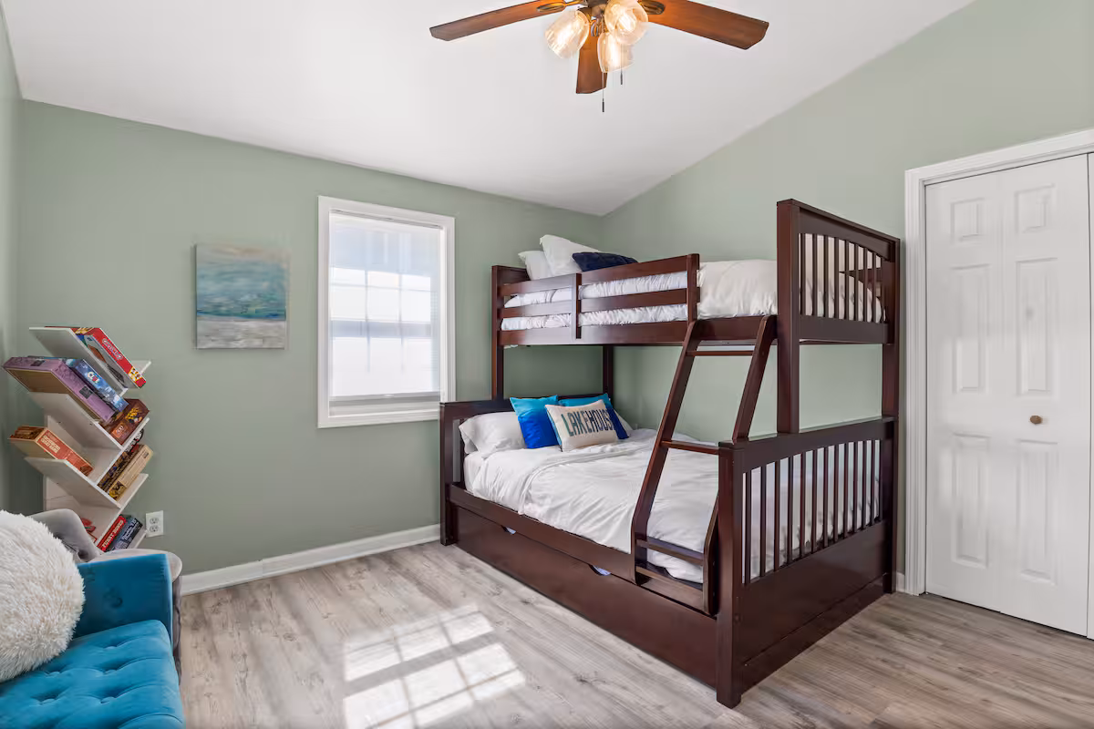 A bedroom in one of our Finger Lakes vacation rentals features a wooden bunk bed, cozy pillows, bookshelf, window, ceiling fan, and soothing light green walls.