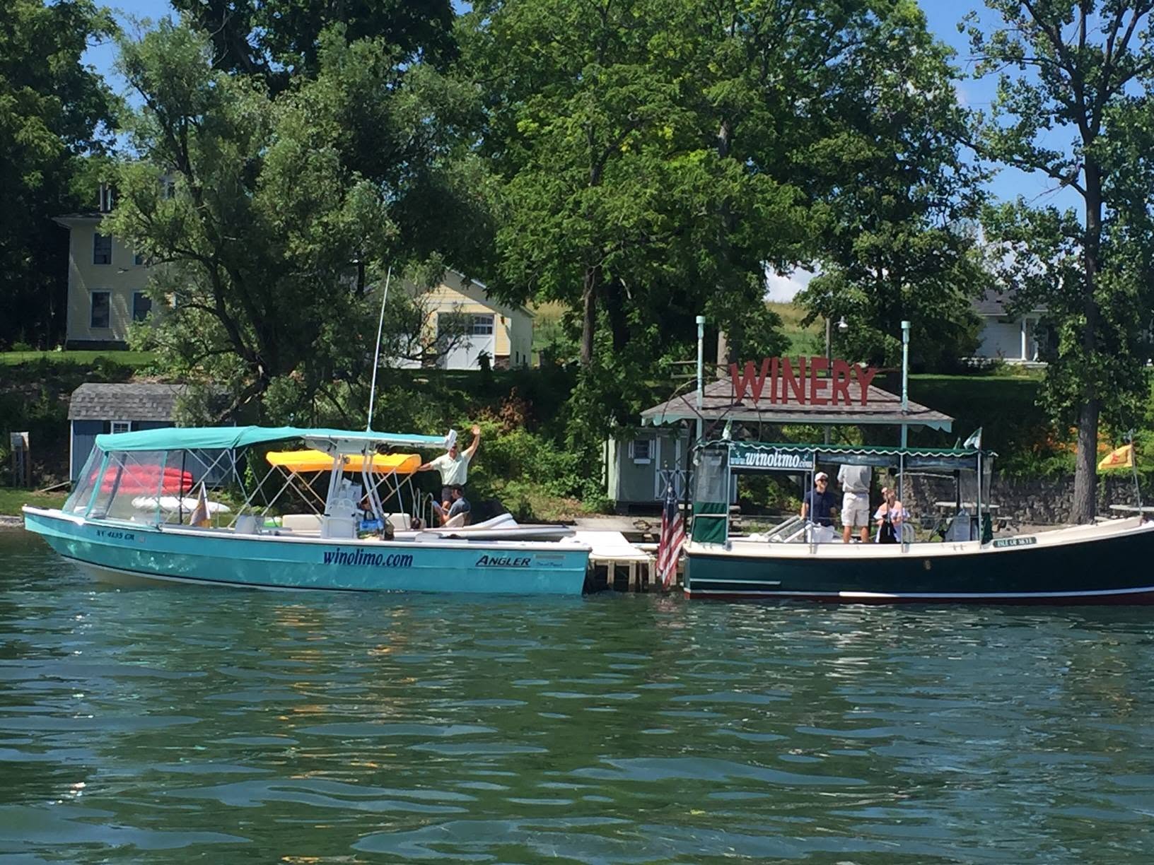 Two boats docked by a lakeside winery, surrounded by trees and houses in the background, make for a perfect stop on Finger Lakes Wine Tours on a sunny day.
