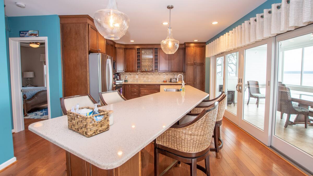 Modern kitchen with wooden cabinets, a large island, wicker chairs, and a view of a sunroom and water outside—perfect for relaxing Finger Lakes vacation rentals or cozy cottages.