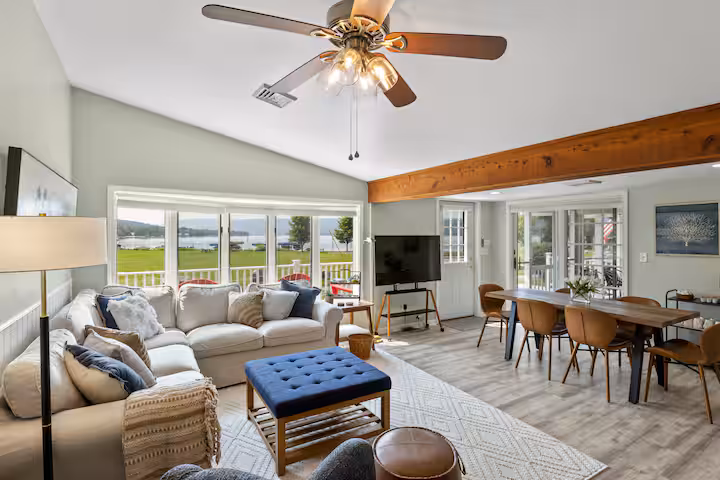 Bright living and dining area with large windows, sectional sofa, TV, and wooden dining table overlooking a green lawn—ideal for relaxing in sought-after Finger Lakes vacation rentals.