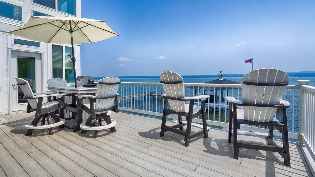 Deck with Adirondack chairs, table, and umbrella overlooking a calm lake and an American flag in the distance—perfect for relaxing at one of the cozy Finger Lakes vacation rentals.
