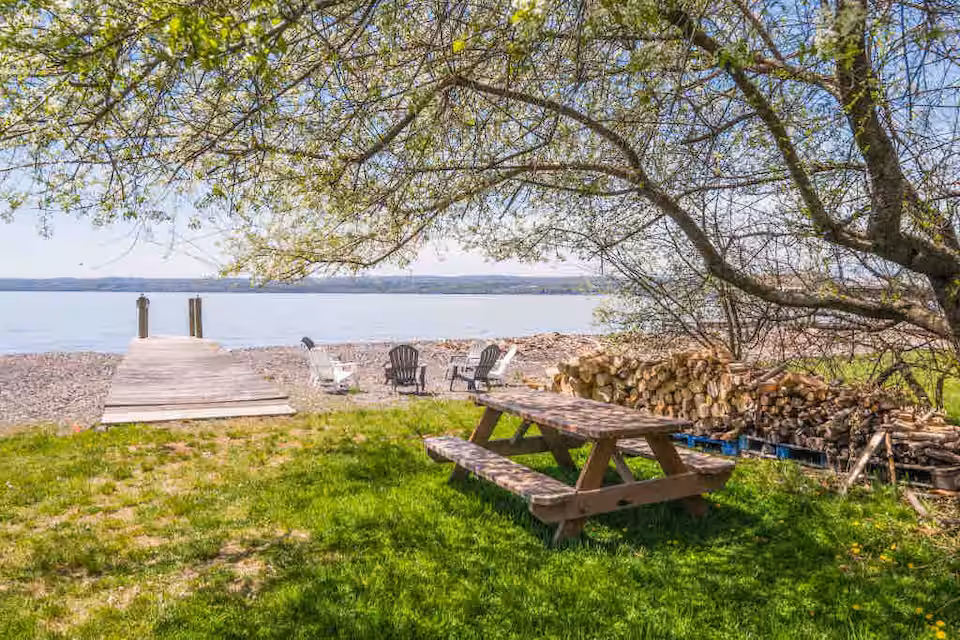 Wooden picnic table and stacked firewood under a tree by a dock and lake with chairs on a sunny day—perfect for relaxing at Finger Lakes vacation rentals or cozy cabins.