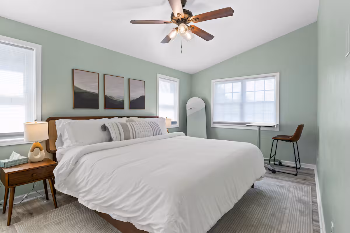 Bedroom with a large bed, green walls, ceiling fan, desk, chair, and three framed pictures above the bed—perfect for relaxing during your stay at one of our Finger Lakes vacation rentals.
