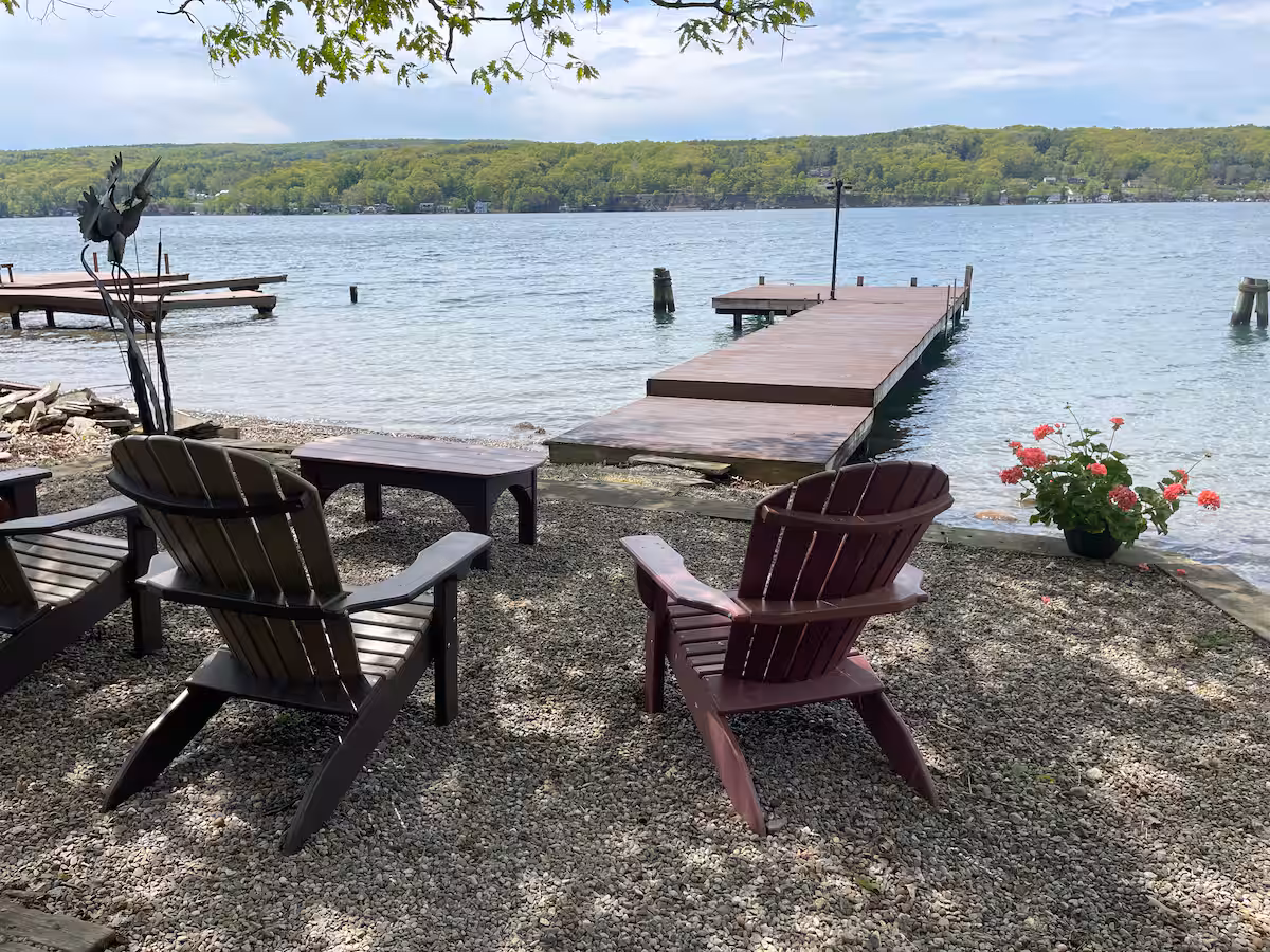 Adirondack chairs face a wooden dock on a calm lake, with flowers and trees in the background—perfect for relaxing at one of the charming Finger Lakes vacation rentals, cottages, or cabins.