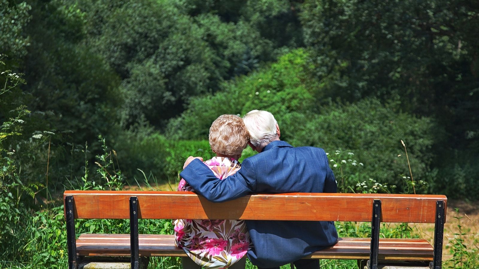 An elderly couple sits close together on a bench, surrounded by green trees and nature, perhaps reflecting on the 9 countries ranked ahead of us for retirement.