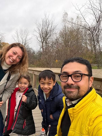 Four people, two adults and two kids, smiling outdoors on a cloudy day with trees and a wooden deck in the background, enjoying their stay at one of the cozy Finger Lakes vacation rentals.