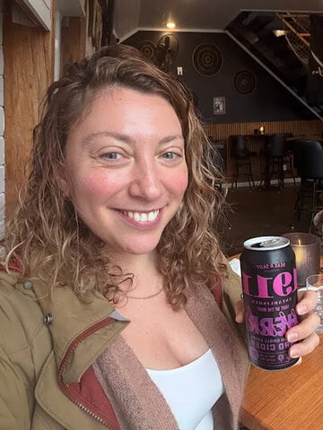 A woman smiles, holding a can, sitting at a wooden table in a cozy café that evokes the charm of Finger Lakes wineries with restaurants; a child sits in the background.