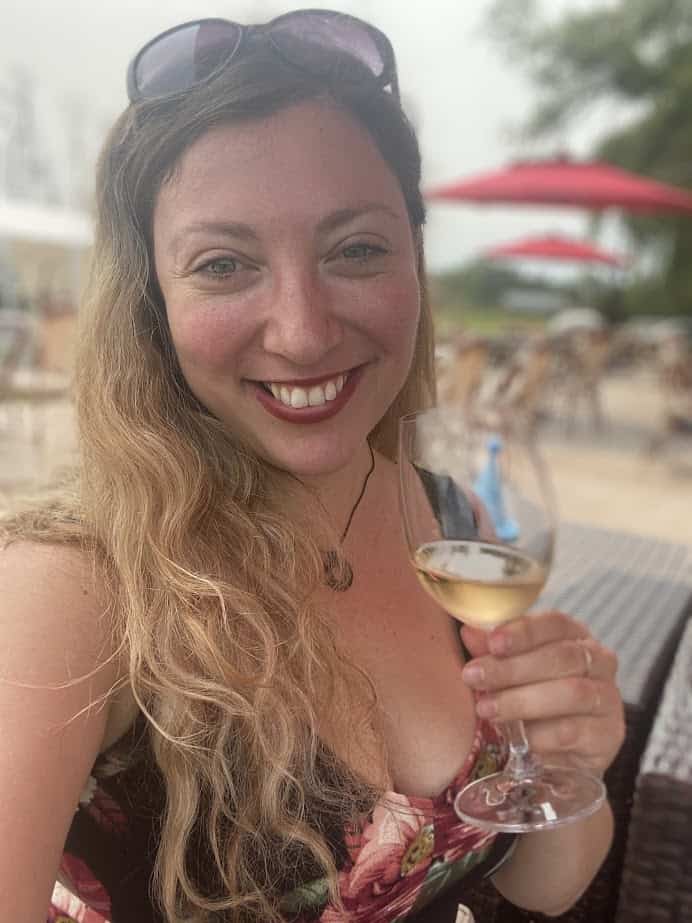 Smiling woman with long blonde hair holding a glass of white wine outdoors at a patio with red umbrellas, enjoying the ambiance of Finger Lakes Wine Tours.