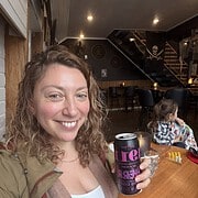 Woman smiling and holding a can in a cozy café, with a child playing at a nearby table—evoking the warm, welcoming vibe found at Finger Lakes wineries with restaurants.