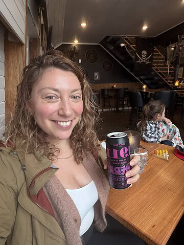 Woman smiling and holding a can in a cozy café, with a child playing at a nearby table—evoking the warm, welcoming vibe found at Finger Lakes wineries with restaurants.