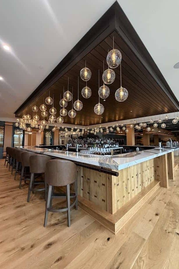 A modern bar with wooden floors, hanging globe lights, and padded barstools around a large marble counter—perfect for unwinding after a day at the Spa or exploring the scenic Finger Lakes.
