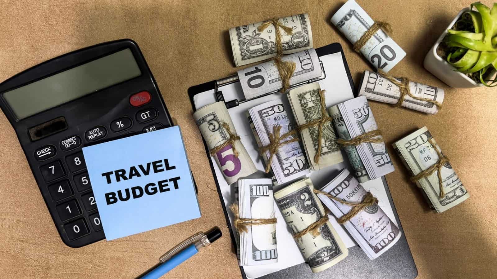 Calculator, clipboard, pen, plant, and bundles of cash with a note reading "TRAVEL BUDGET" on a desk.