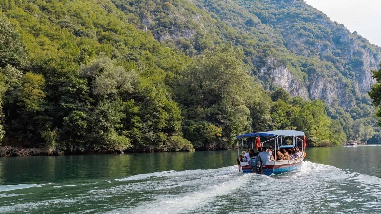 A boat with people cruises on a river surrounded by lush green hills and dense forest, reminiscent of hidden European destinations cost less than you might expect.