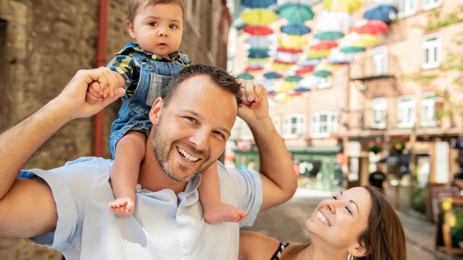 A smiling man carries a baby on his shoulders while a woman smiles at them; colorful umbrellas hang above.