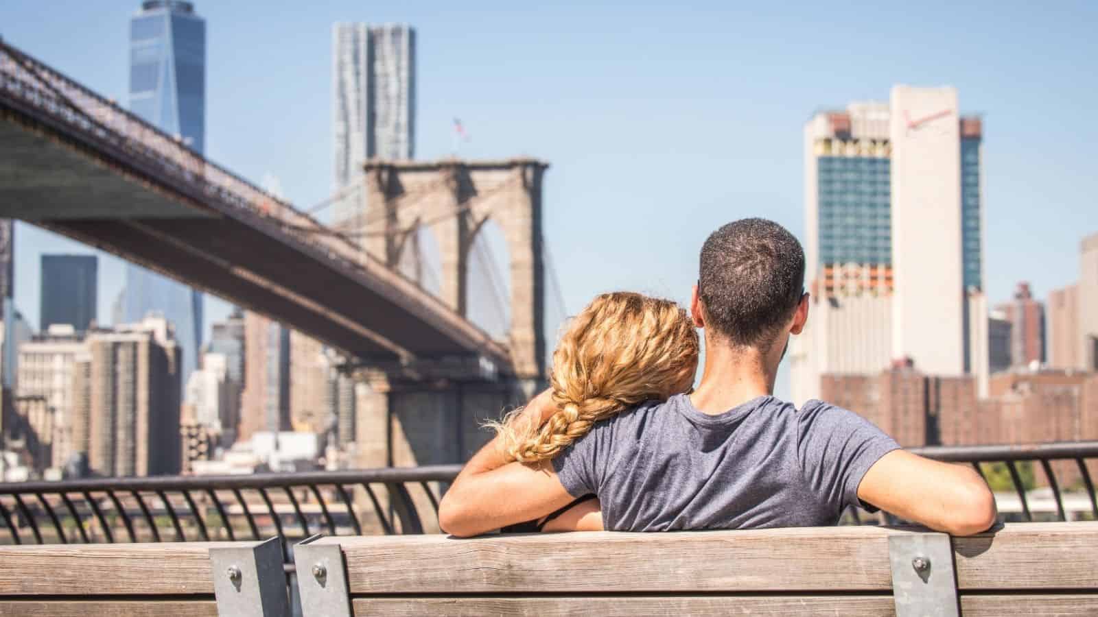 A couple sits on a bench, facing the Brooklyn Bridge and Manhattan skyline on a sunny day.